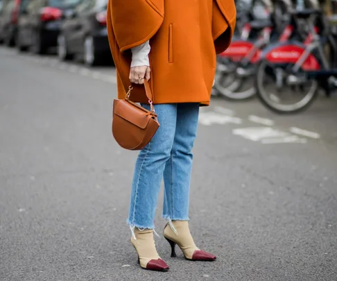 Person wearing an orange coat, holding a brown handbag, with jeans and high-heeled shoes on a city street.