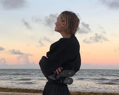 Person in black attire stands on beach at sunset with ocean in background, holding shoes behind their back.