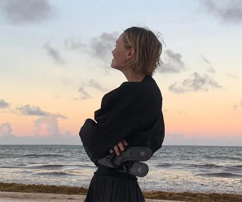 Person in black attire stands on beach at sunset with ocean in background, holding shoes behind their back.