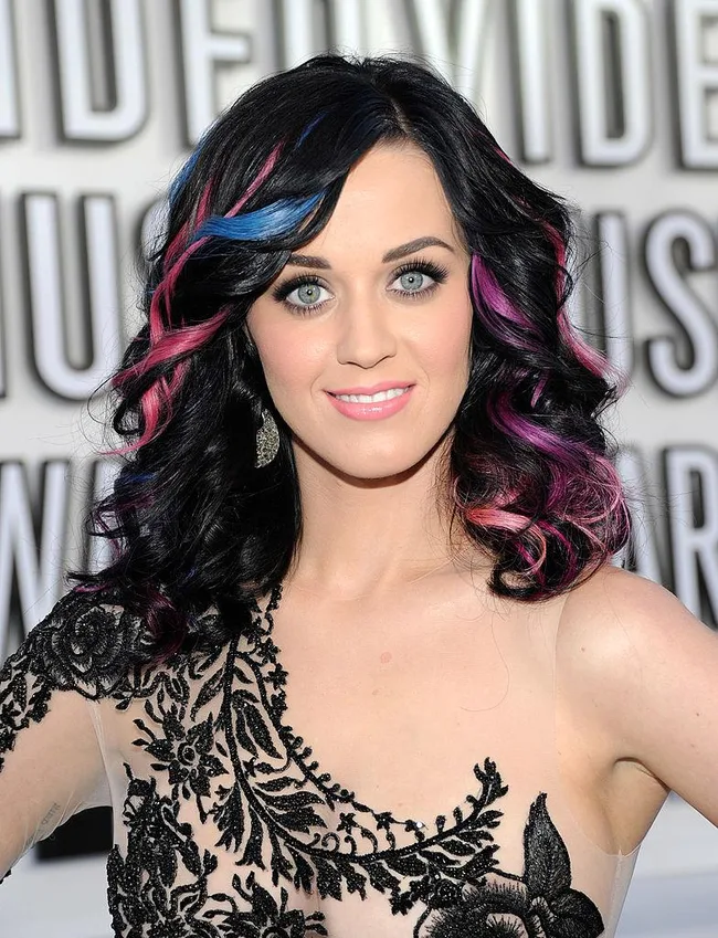 A woman with colorful highlights in wavy hair, wearing an ornate black and nude dress, smiles at a formal event.