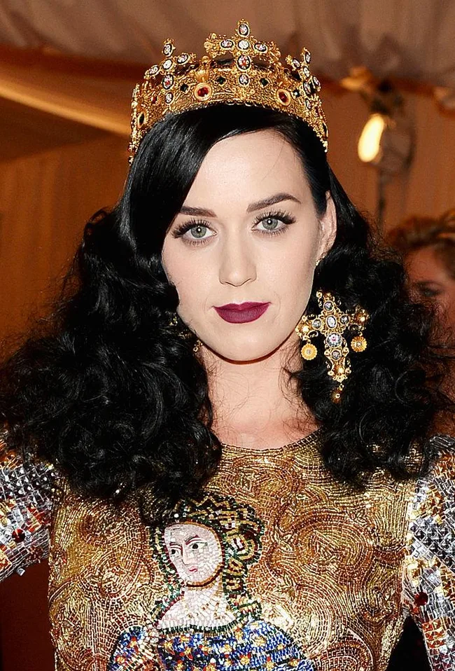 A person with long curly black hair wears an ornate gold crown and a detailed, colorful outfit with a regal design.