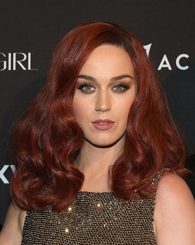 Red-haired woman in sparkly dress at an event, with wavy hair and nose ring, posing against a dark background.