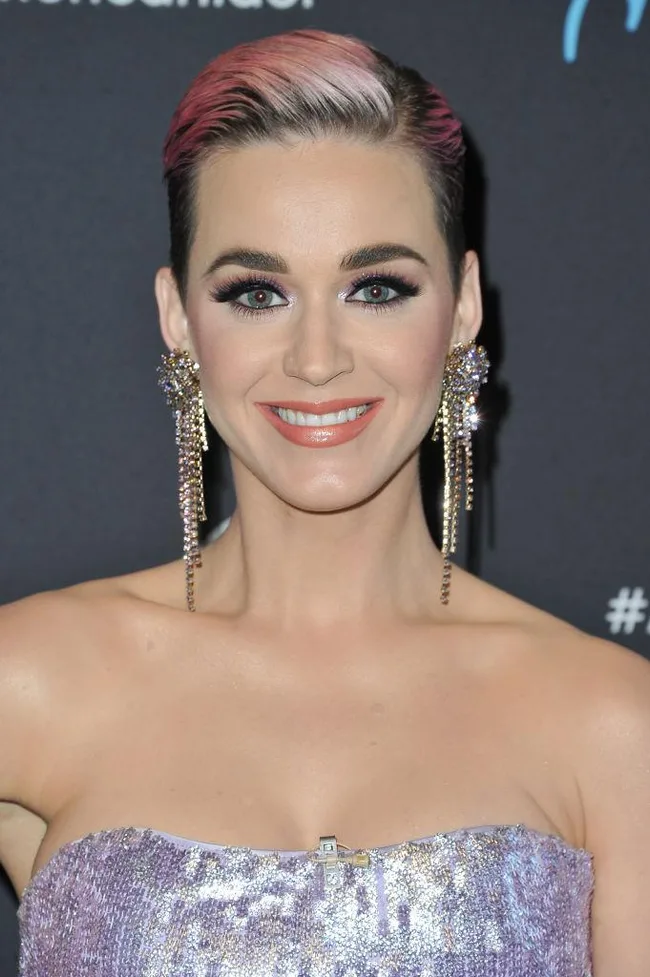 A person with short pink-highlighted hair and sparkling earrings smiles at an event, wearing a strapless sequin top.