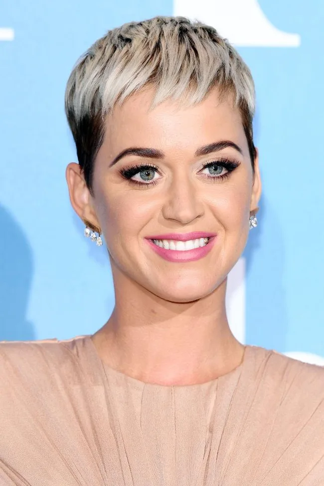 A person with short blonde hair smiling, wearing a beige top against a blue background.