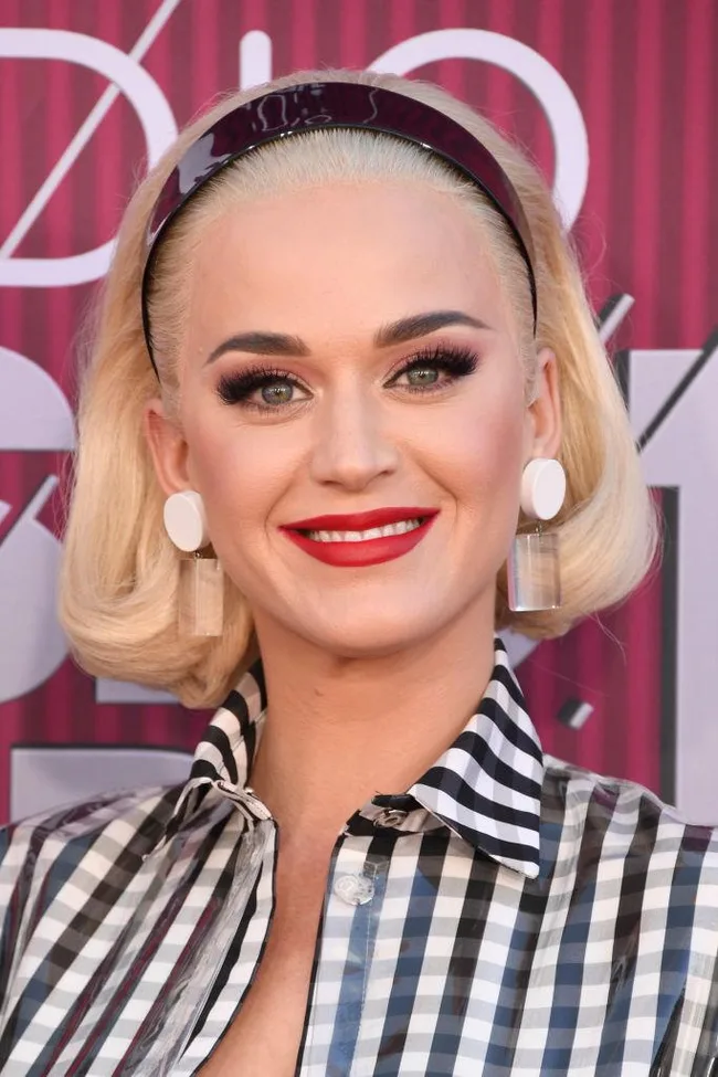 Smiling woman with blond hair, wearing large earrings and a checkered top, against a red and purple striped background.