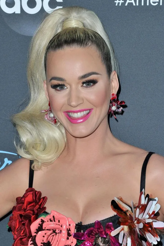 A person with blonde hair in ponytail, wearing floral dress and pink lipstick, smiling at an event.