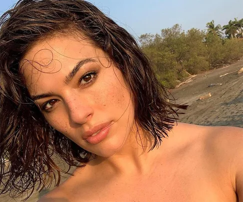 Woman with wet hair and natural makeup taking a selfie on a beach.