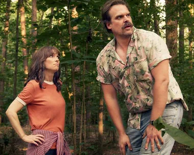 A man and woman in a forest; the man looks alert. They're dressed in casual clothing.