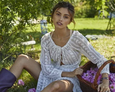 Woman in a white dress sitting on grass with a basket of flowers, surrounded by greenery.