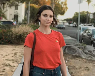 A woman in a red t-shirt stands on a suburban street, with a slight smile and a backpack over her shoulder.