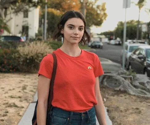 A woman in a red t-shirt stands on a suburban street, with a slight smile and a backpack over her shoulder.