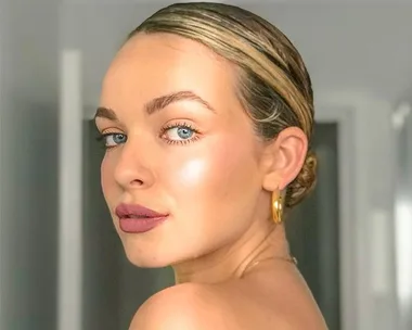 Close-up of a woman with slicked-back hair, gold hoop earrings, and glowing makeup, looking over her shoulder.