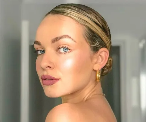 Close-up of a woman with slicked-back hair, gold hoop earrings, and glowing makeup, looking over her shoulder.