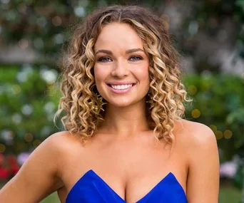 A woman with curly hair in a blue dress, smiling outdoors with a blurred garden background.