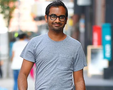 A person with glasses and a gray t-shirt walks in an urban setting, smiling.