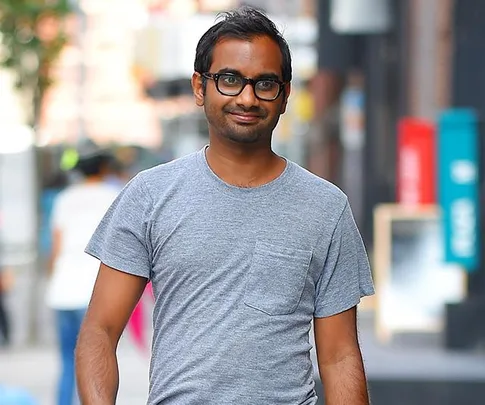 A person with glasses and a gray t-shirt walks in an urban setting, smiling.