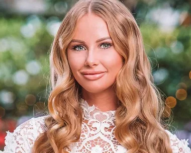 Blonde woman with wavy hair and a lace dress smiles outdoors with a blurred green background.