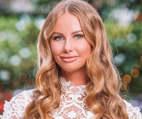 Blonde woman with wavy hair and a lace dress smiles outdoors with a blurred green background.