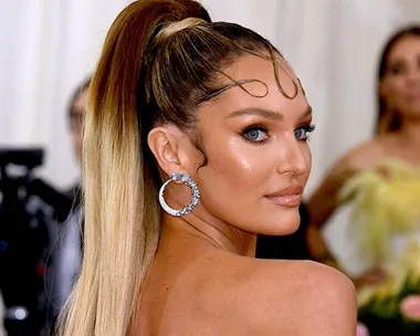 Model with high ponytail, wavy hair strands on forehead, silver hoop earrings, and glowing makeup on a red carpet event.