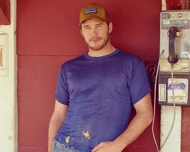 Man in a blue T-shirt and cap leaning against a red wall next to a vintage payphone.