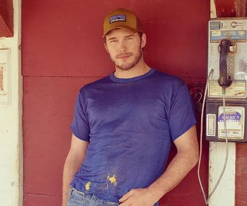 Man in a blue T-shirt and cap leaning against a red wall next to a vintage payphone.