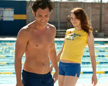 Two people smiling by a swimming pool; one is shirtless, the other in a yellow sports top and blue shorts.