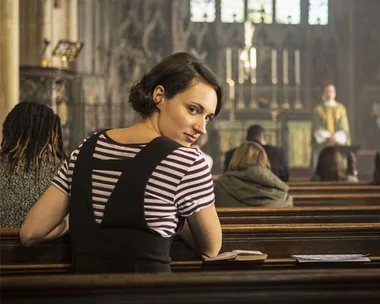 Woman in striped shirt turns to look back while seated in a church pew, with a blurred priest in the background.