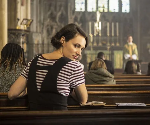 Woman in striped shirt turns to look back while seated in a church pew, with a blurred priest in the background.