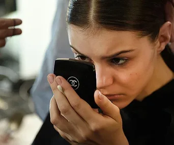 Woman closely inspecting her face in a small Chanel mirror, possibly looking at acne scars.