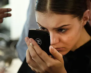 Woman closely inspecting her face in a small Chanel mirror, possibly looking at acne scars.