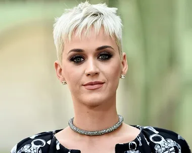 A person with short blonde hair, wearing a patterned outfit and a sparkly necklace, outdoors with blurred background.