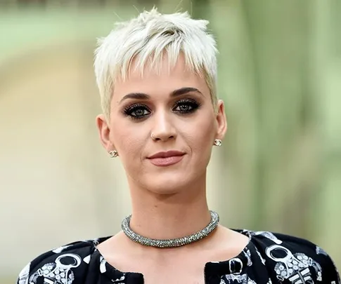 A person with short blonde hair, wearing a patterned outfit and a sparkly necklace, outdoors with blurred background.