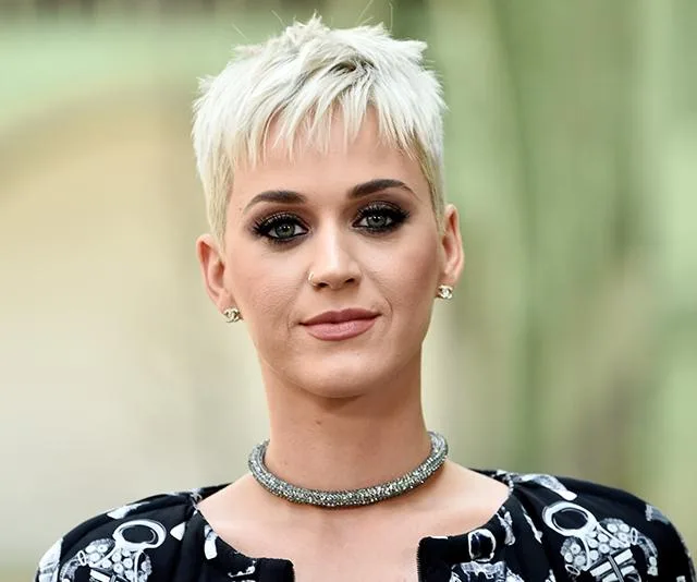 A person with short blonde hair, wearing a patterned outfit and a sparkly necklace, outdoors with blurred background.