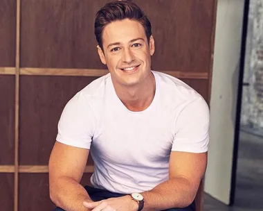 Man in a white t-shirt smiling, sitting indoors with wood panel wall background.