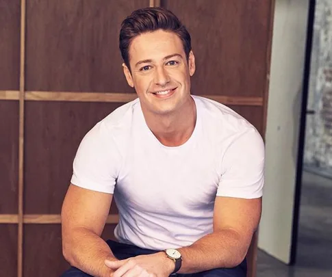 Man in a white t-shirt smiling, sitting indoors with wood panel wall background.