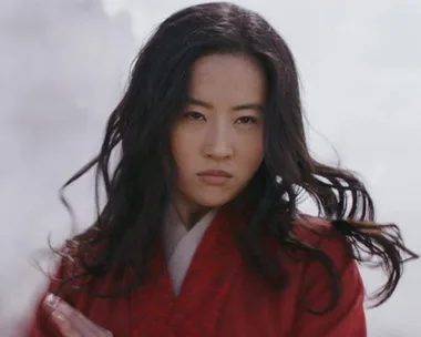 Mulan from live-action film, in red robe, looking determined with flowing hair against a cloudy backdrop.