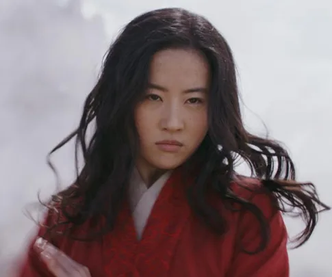 Mulan from live-action film, in red robe, looking determined with flowing hair against a cloudy backdrop.