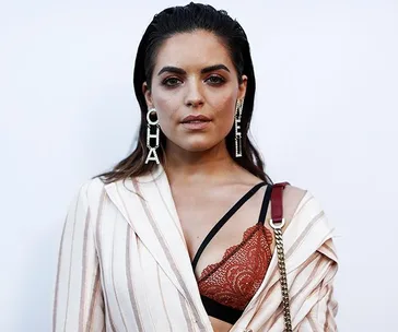 Woman in striped blazer with bold earrings and lace bralette, looking directly at camera against plain background.