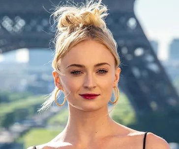 Blonde woman with updo hairstyle and hoop earrings, standing in front of the Eiffel Tower during day.