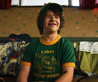 Dustin from Stranger Things in a green "Camp Know Where '85” shirt, smiling in a retro-themed room.