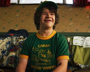 Dustin from Stranger Things in a green "Camp Know Where '85” shirt, smiling in a retro-themed room.