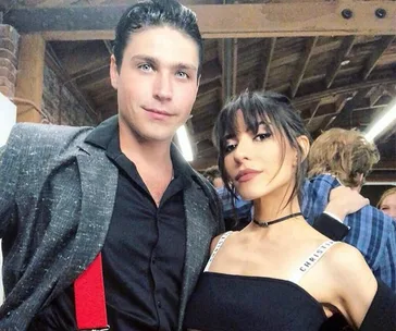 A man in a black shirt and gray jacket with red suspenders poses with a woman in a black top under wooden beams.