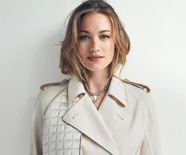 A woman with wavy hair in a beige trench coat and necklace, standing against a plain background.