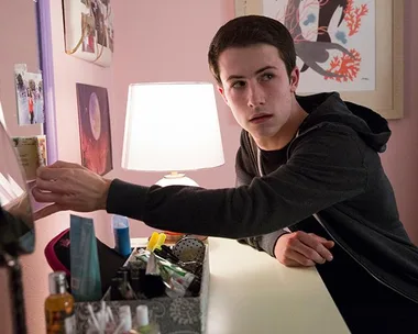 A teenage boy reaches for a photo on a desk in a dimly lit room, with a lamp and various items around.