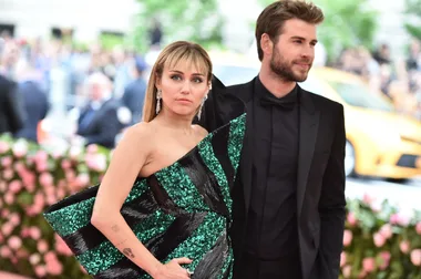 A couple poses at a glamorous event, with a background of greenery and flowers.