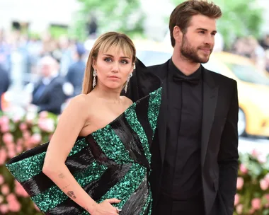 A couple poses at a glamorous event, with a background of greenery and flowers.