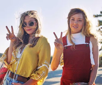 Two women pose outdoors, making peace signs, one in a yellow top and sunglasses, the other in red overalls.