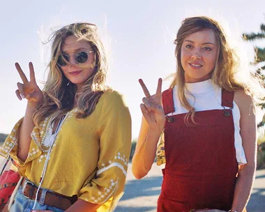 Two women pose outdoors, making peace signs, one in a yellow top and sunglasses, the other in red overalls.