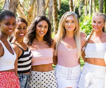 Group of five smiling women in summer outfits standing together with trees in the background.