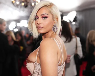 A woman with blonde hair and pink dress posing on a red carpet, with chandelier and crowd in the background.